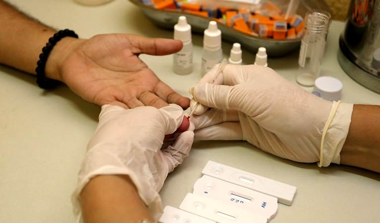 a foto mostra as mãos de um enfermeiro, de luvas, usando uma agulha no dedo de uma pessoa para fazer o teste de HIV, sífilis e hepatite